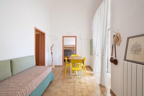 Ein schmaler Raum mit einem Bett, einem Tisch mit gelben Stühlen, einer Holztür und einem gerahmten Bild an der Wand.