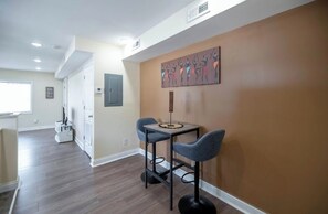 A modern kitchen with a dining area, featuring a kitchen island with bar stools, a wall-mounted artwork, and a window providing natural light. - Queen City Hub (Cincinnati)
