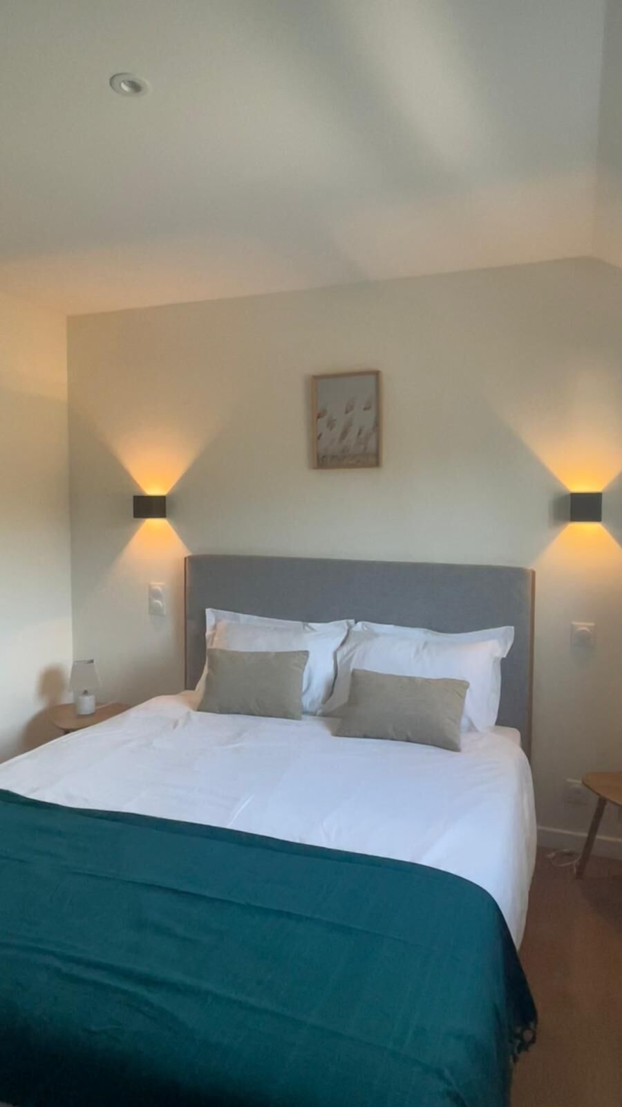 A hotel room with a bed, a headboard, two wall-mounted lights, a painting, and a small table with a lamp.