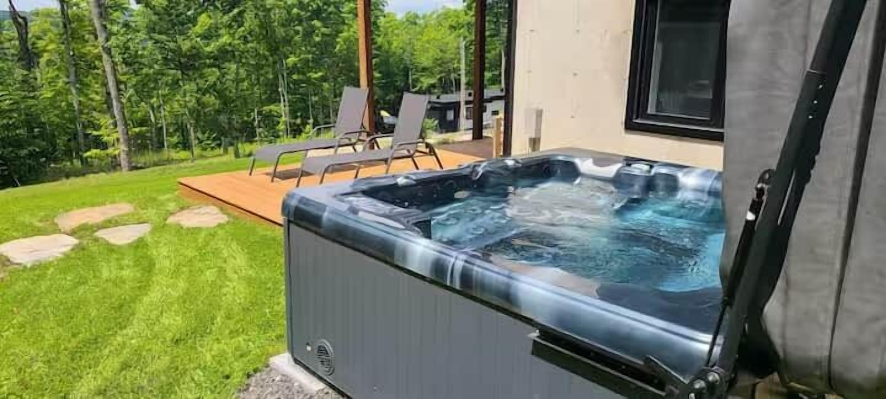Een hottub in een achtertuin met een houten terras, gazon en een raam met uitzicht op de omgeving.