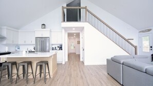 A modern kitchen with a central island, bar stools, a staircase, and a grey sofa.