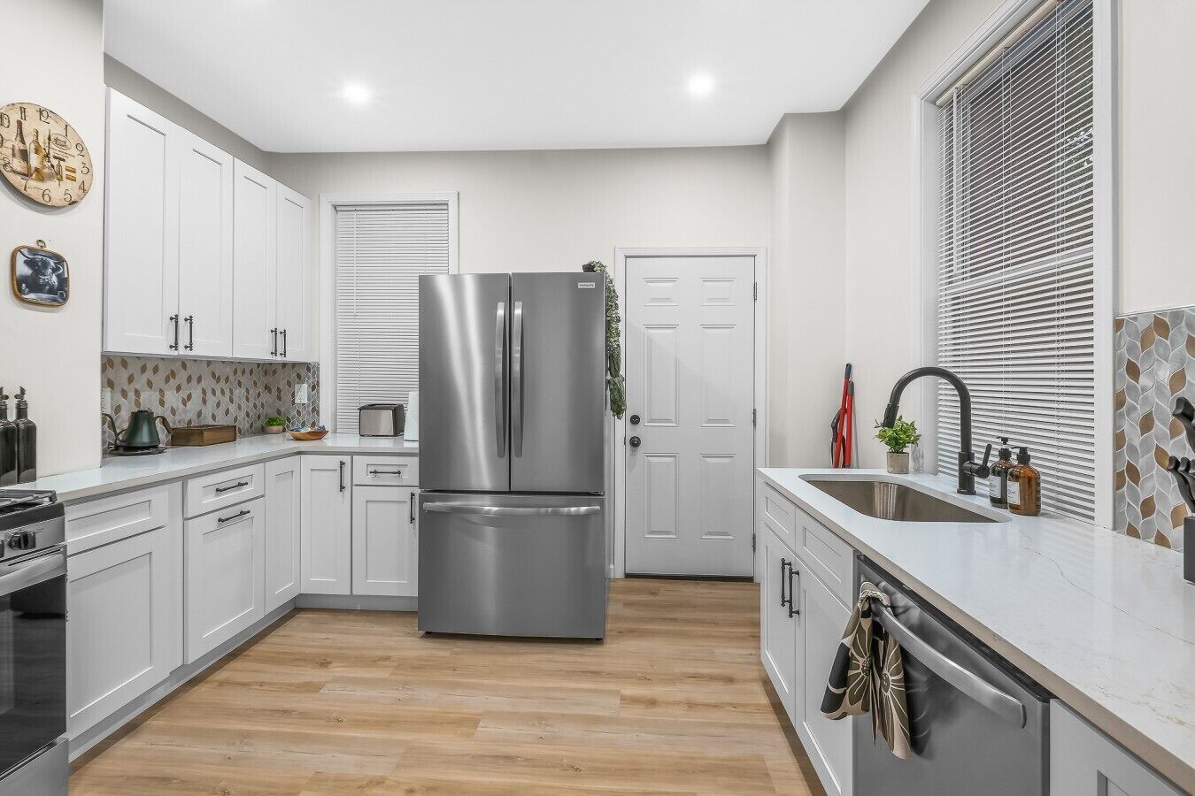 A modern kitchen with white cabinets, a stainless steel refrigerator, wooden flooring, and a window with blinds.