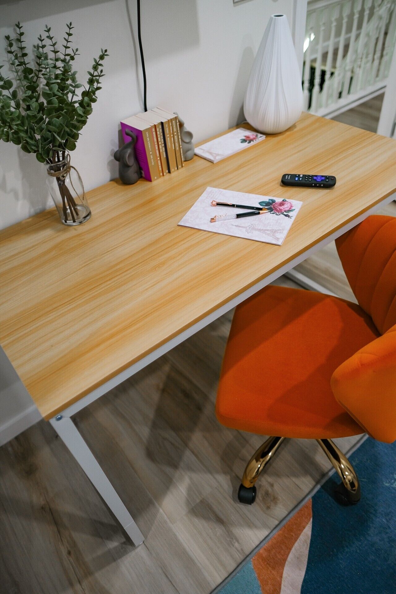 A wooden desk with a vase of green plants, a remote control, and a pen on a notepad.