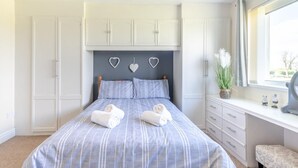 Ein Schlafzimmer mit einem Bett, zwei weißen Handtüchern, einem Schminktisch und einem Fenster mit Vorhängen, an einer Wand befinden sich herzförmige Verzierungen.