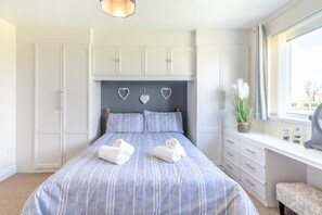 Ein Schlafzimmer mit einem Bett, zwei weißen Handtüchern, einem Schminktisch und einem Fenster mit Vorhängen, an einer Wand befinden sich herzförmige Verzierungen.