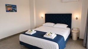 Ein Hotelzimmer mit einem großen Bett, weißen Bettwäsche, einem dunkelblauen Kopfteil, zwei Wandleuchten, einer Klimaanlage und einem Gemälde an der Wand.