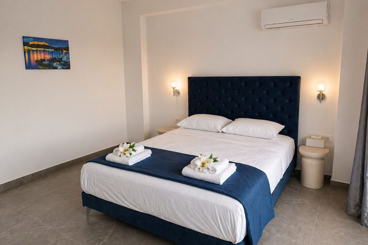 Ein Hotelzimmer mit einem großen Bett, weißen Bettwäsche, einem dunkelblauen Kopfteil, zwei Wandleuchten, einer Klimaanlage und einem Gemälde an der Wand.