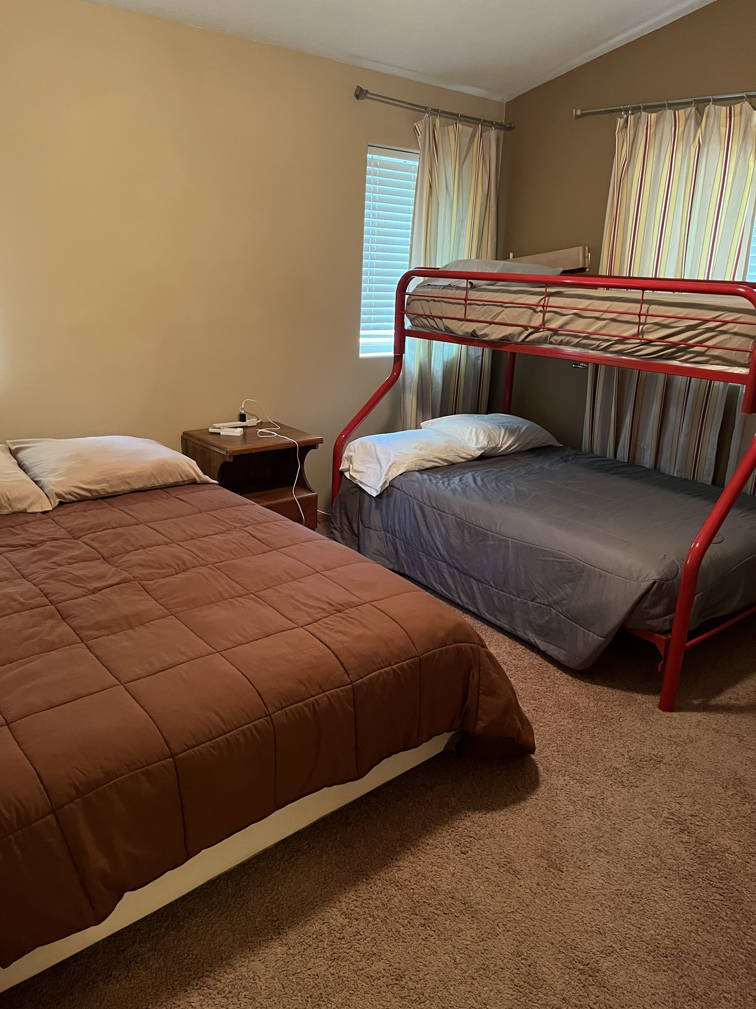 A room with two beds, one with a red frame and one with a brown frame, a nightstand with a lamp, and a window with curtains.