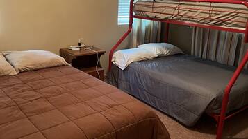 A room with two beds, one with a red frame and one with a brown frame, a nightstand with a lamp, and a window with curtains.