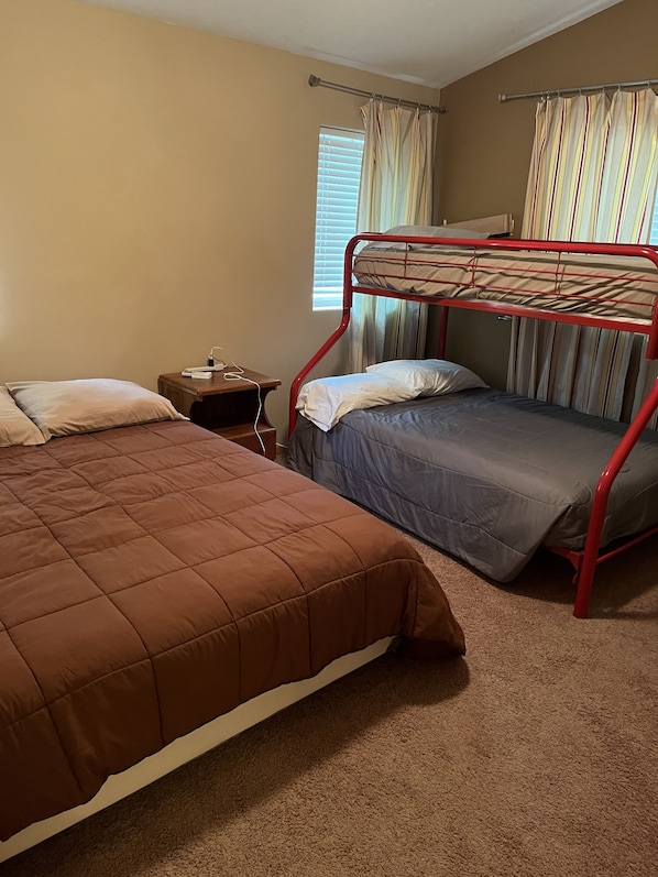 A room with two beds, one with a red frame and one with a brown frame, a nightstand with a lamp, and a window with curtains.