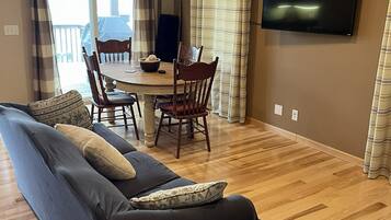 A living room with a sofa, a dining table with chairs, a television mounted on the wall, and a wooden floor.