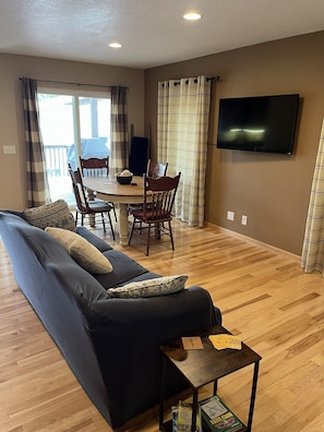A living room with a sofa, a dining table with chairs, a television mounted on the wall, and a wooden floor.