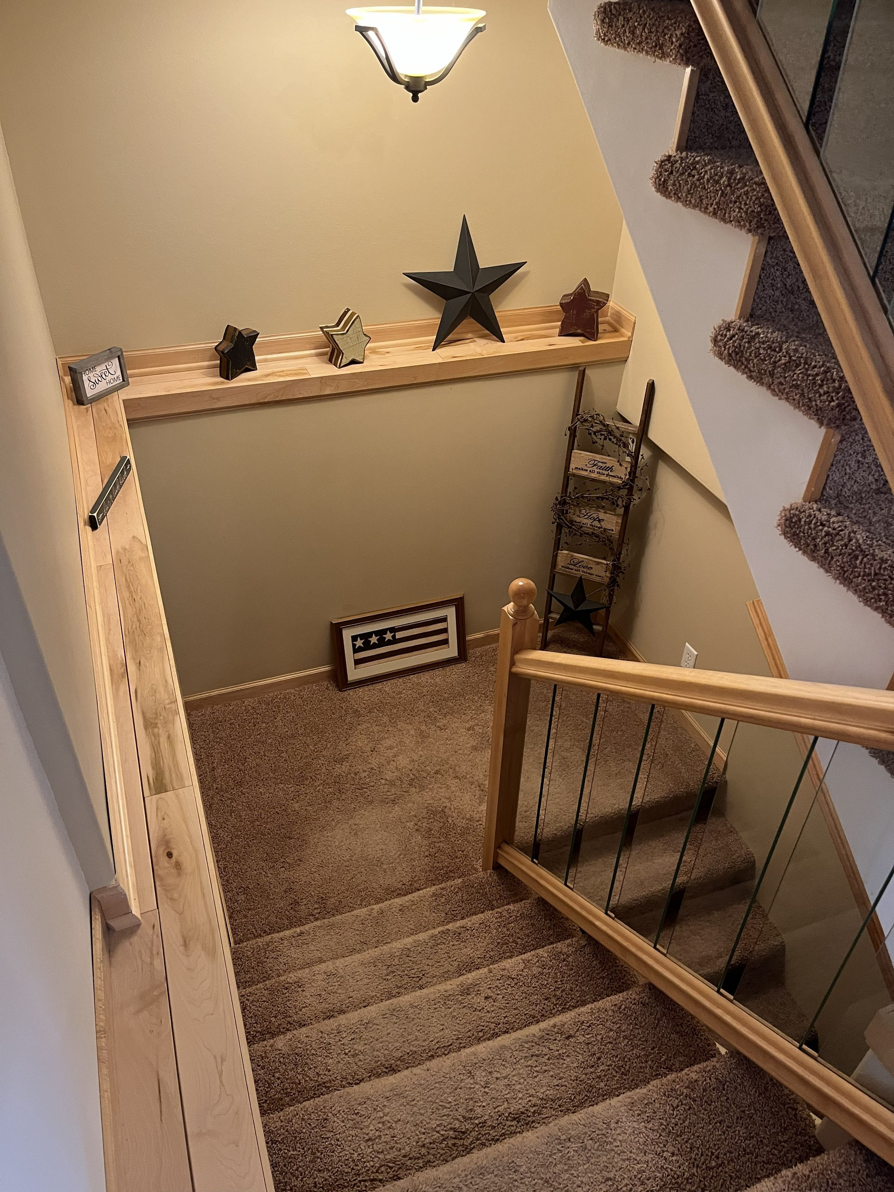 A carpeted staircase with a wooden handrail and glass balusters, leading to a landing with a shelf holding decorative items and a framed sign.