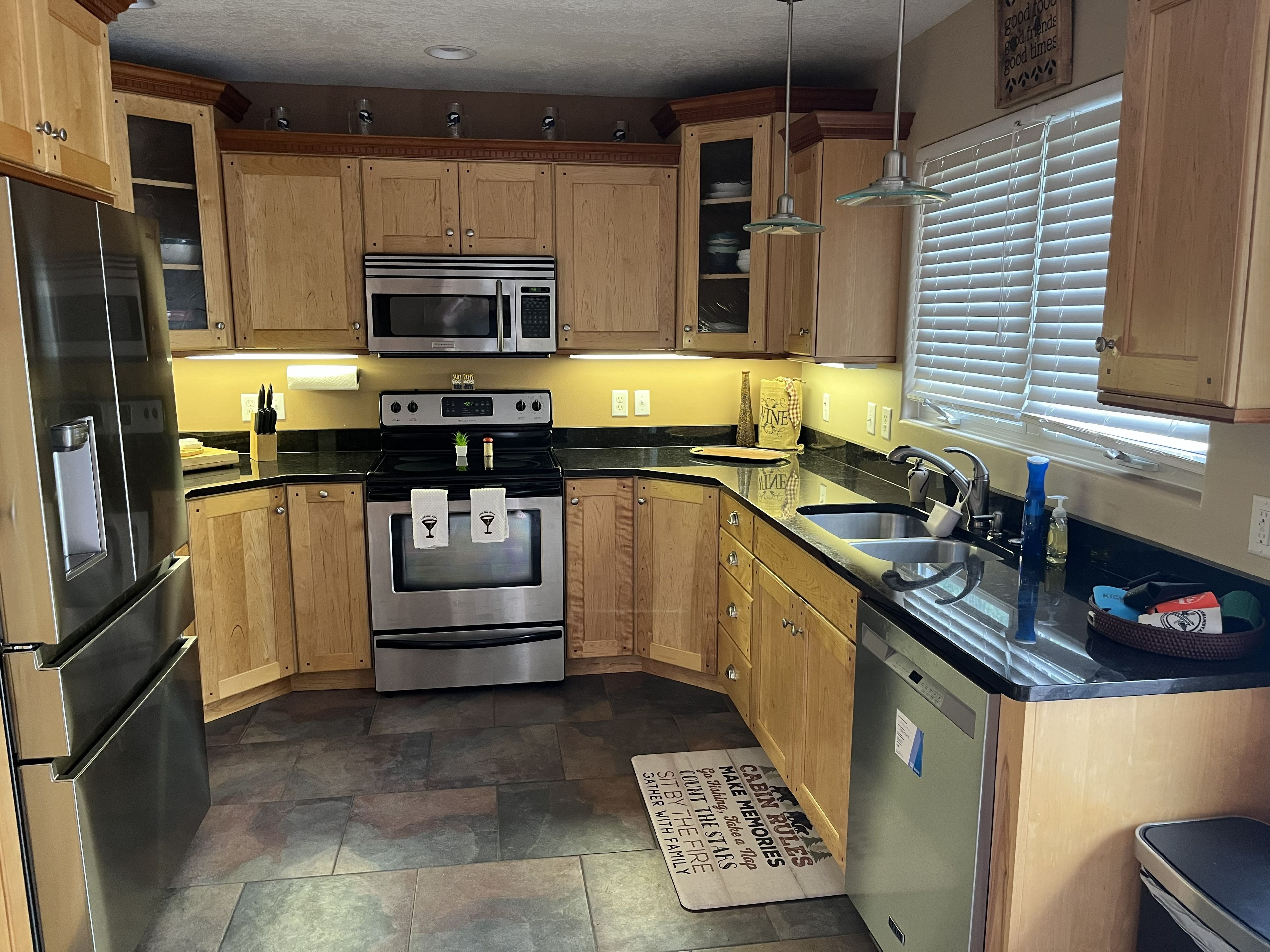 A kitchen with wooden cabinets, stainless steel appliances, a black countertop, and a window with blinds.