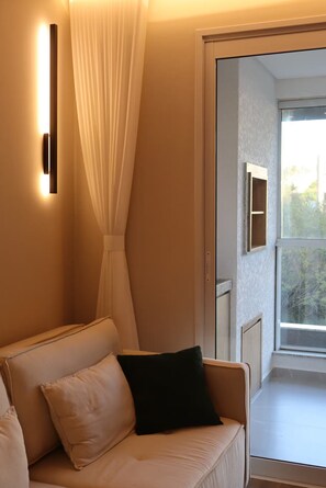 A modern room with a sofa, a wall-mounted light, a sheer curtain, and a view through a window. - New and cozy: on the beach and with a pool (Ingleses, Florianópolis) (Florianópolis)