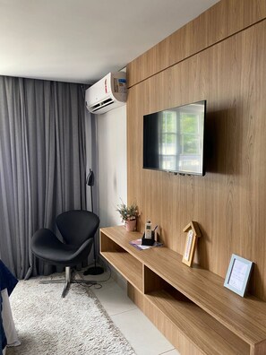 A modern room with a mounted TV, a wooden console, a black chair, a potted plant, and an air conditioning unit. - STÚDIO GRANJA BRASIL - APTO 3-414 (Petrópolis)