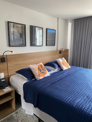 A hotel room with a bed, a nightstand, a lamp, and a window with curtains. - STÚDIO GRANJA BRASIL - APTO 3-414 (Petrópolis)
