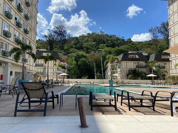 A hotel pool area with lounge chairs and umbrellas, surrounded by buildings and a hillside.