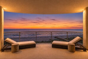 Ein Balkon mit zwei gepolsterten Stühlen und einem kleinen runden Tisch, mit Blick auf einen Sonnenuntergang.