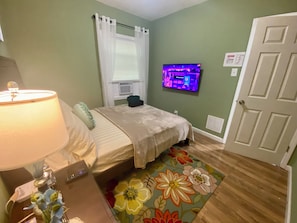 Ein Schlafzimmer mit einem Bett, einem an der Wand montierten Fernseher, einer Tür und einem Fenster mit Vorhängen.