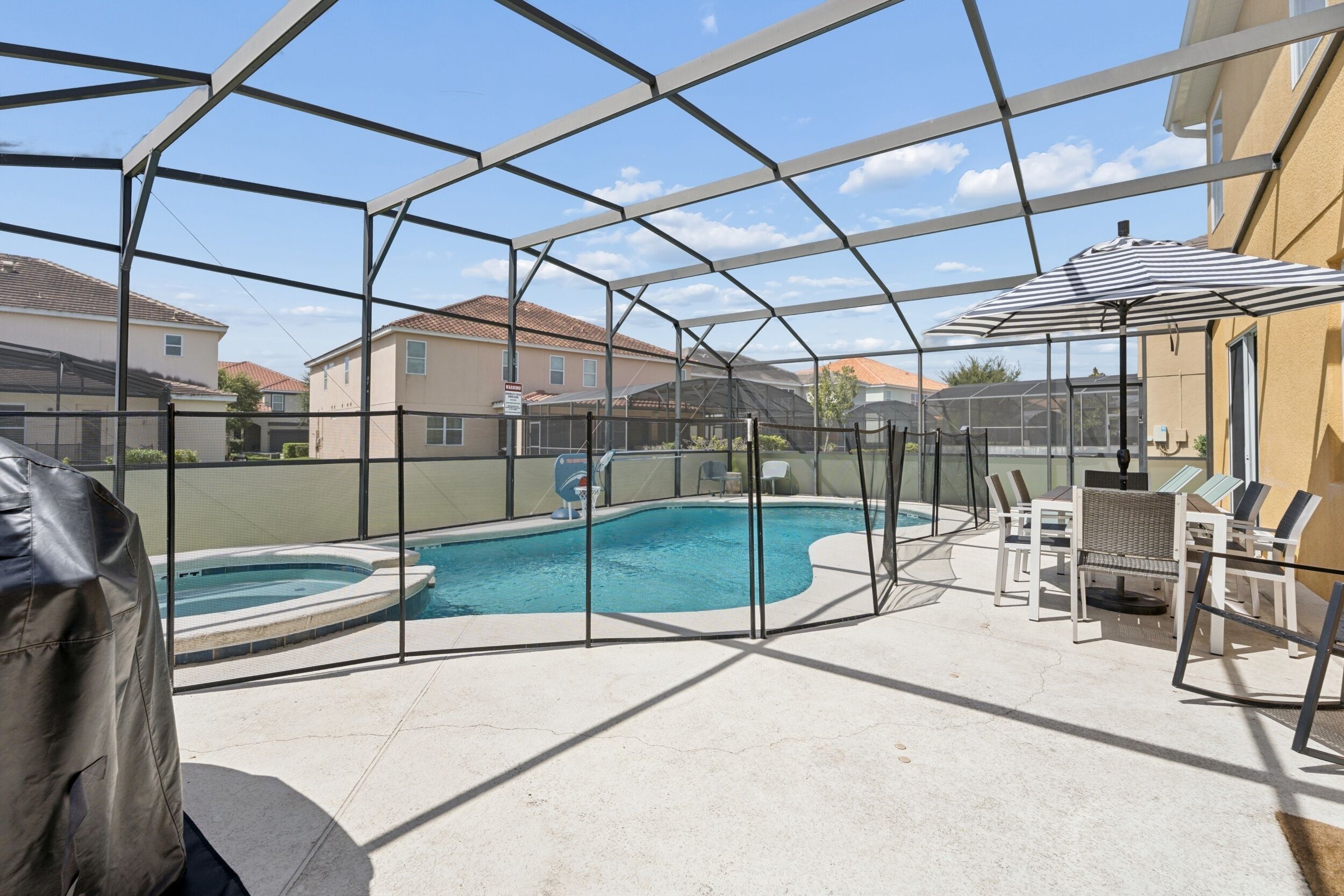 Ein überdachter Außenpool mit Whirlpool, umgeben von einem Metallzaun, und eine Terrassenfläche mit Stühlen und Sonnenschirm.