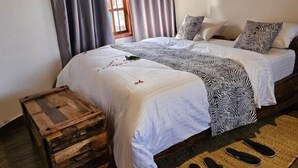 A double bed with a white and black patterned bedspread, two pillows, a wooden chest of drawers, and a woven rug on a wooden floor.