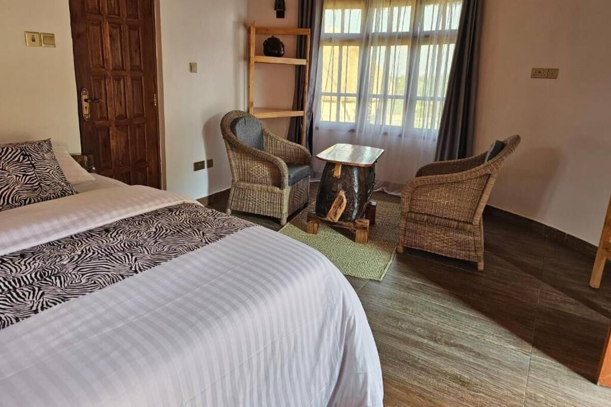 Una stanza con un letto, due poltrone in vimini, un tavolino, una mensola in legno e una finestra con tende.