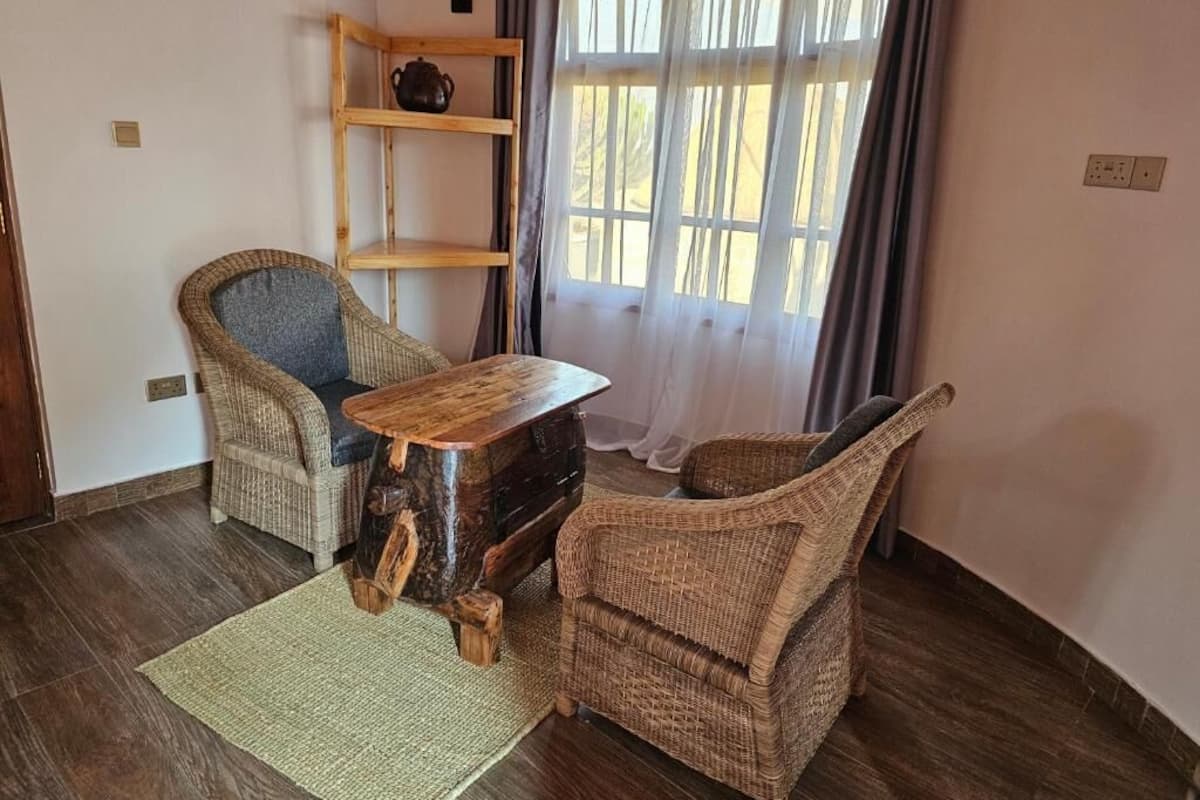 Una stanza con un tavolo in legno, due sedie in vimini, un tappeto intrecciato, una mensola in legno con una pianta e una finestra con tende.
