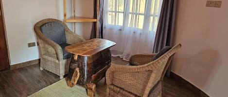 A room with a wooden table, two wicker chairs, a woven rug, a wooden shelf with a pot, and a window with curtains.