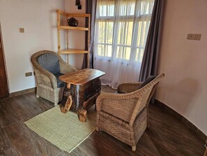 Una habitación con una mesa de madera, dos sillas de mimbre, un tapete tejido, un estante de madera con una maceta y una ventana con cortinas.