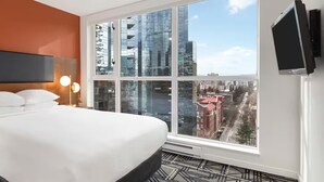 Habitación de hotel con cama, lámparas de noche, televisor de pantalla plana y un ventanal con vista a la ciudad.
