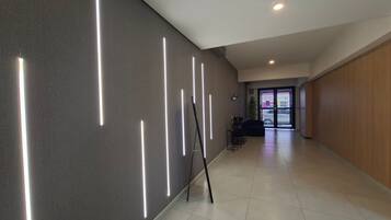 Um corredor com design moderno, piso claro e uma parede com fitas verticais de iluminação. Há um espaço de estar com uma mesa e cadeiras, e uma porta que dá para outro quarto.