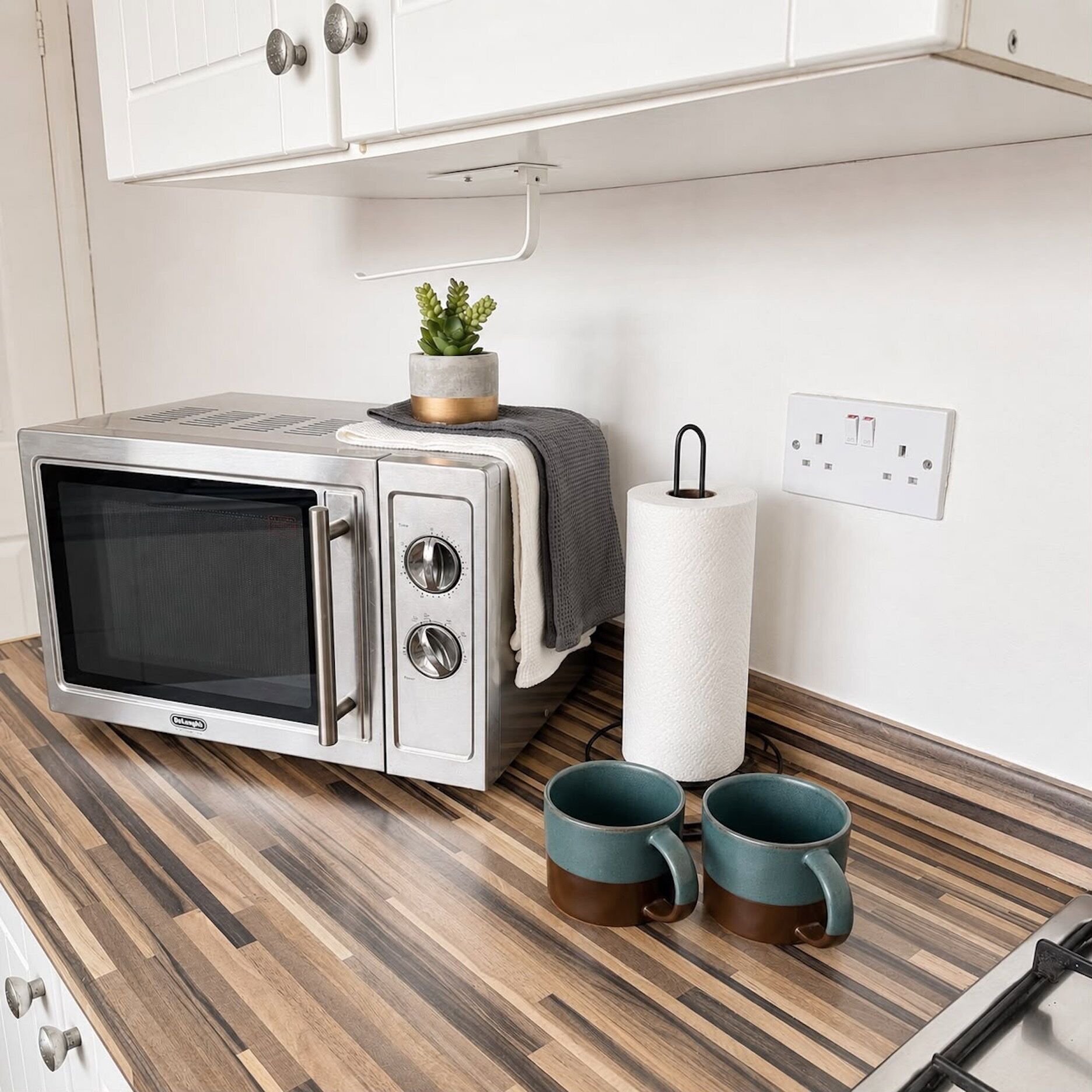 Une petite cuisine équipée d’un micro-ondes, d’une petite plante en pot, d’un rouleau d’essuie-tout et de deux tasses de couleur turquoise posées sur un plan de travail en bois.