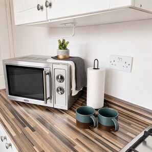 A compact kitchenette with a microwave, a small potted plant, a roll of paper towels, and two teal mugs on a wooden countertop. - 3-Bed Contractor Stay | Comfort & Space (South Shields)
