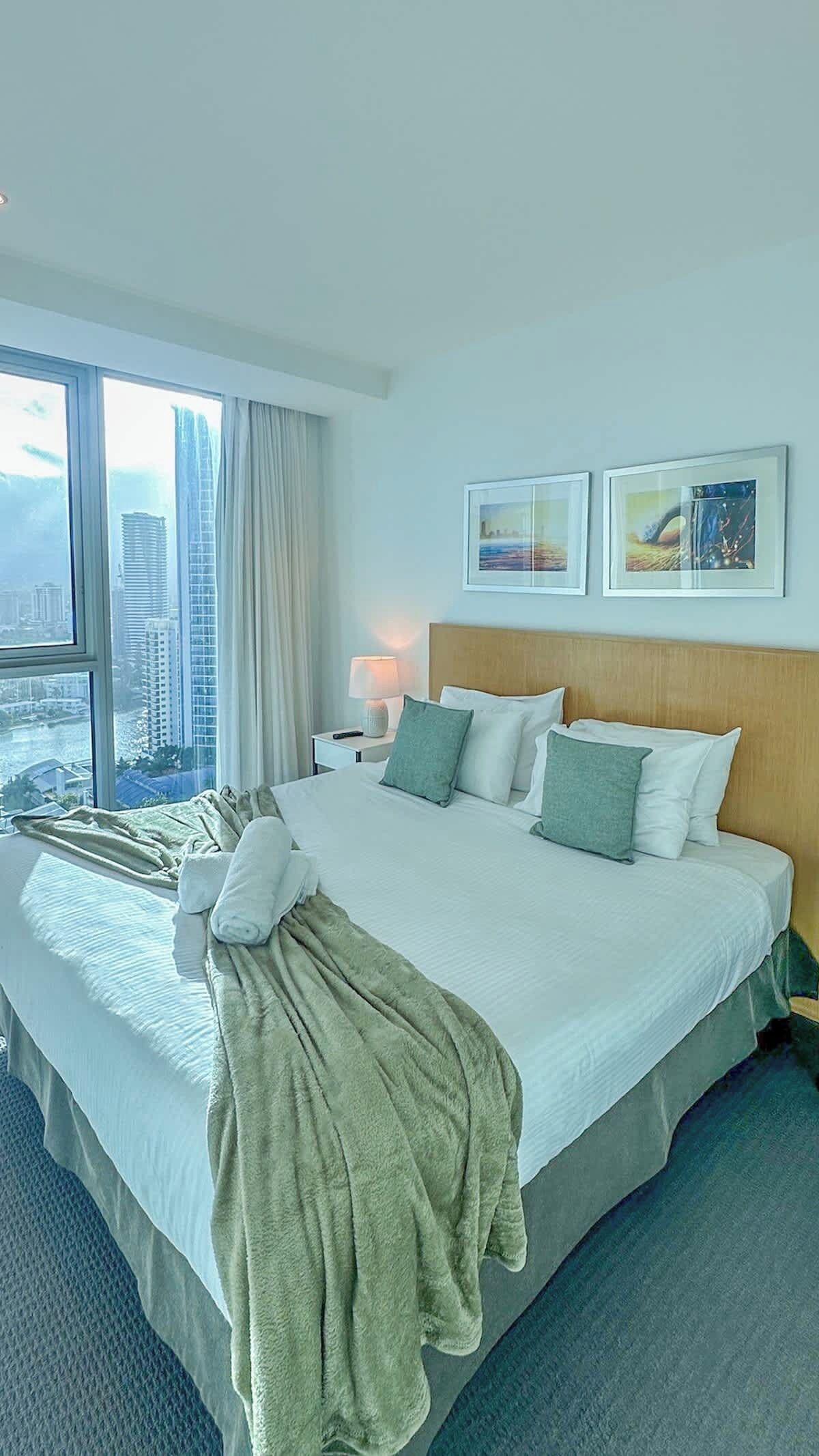 A hotel room with a large bed, a nightstand, a lamp, two framed pictures on the wall, and a view of the city through large windows.