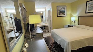 Habitación de hotel con cama, escritorio con lámpara, espejo, baño con ducha y un cuadro en la pared.