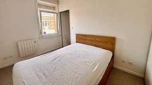 Een slaapkamer met een enkel bed, een radiator, een raam met uitzicht op een gebouw en een deur.