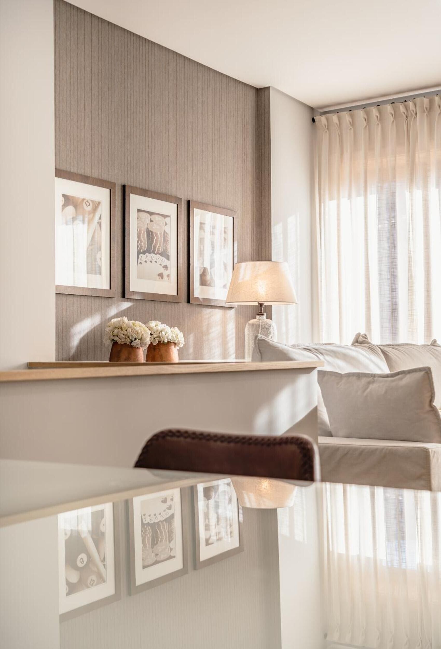 A modern hotel room with a bed, a bedside table with a lamp, a wall-mounted shelf with framed pictures, and a window with sheer curtains.