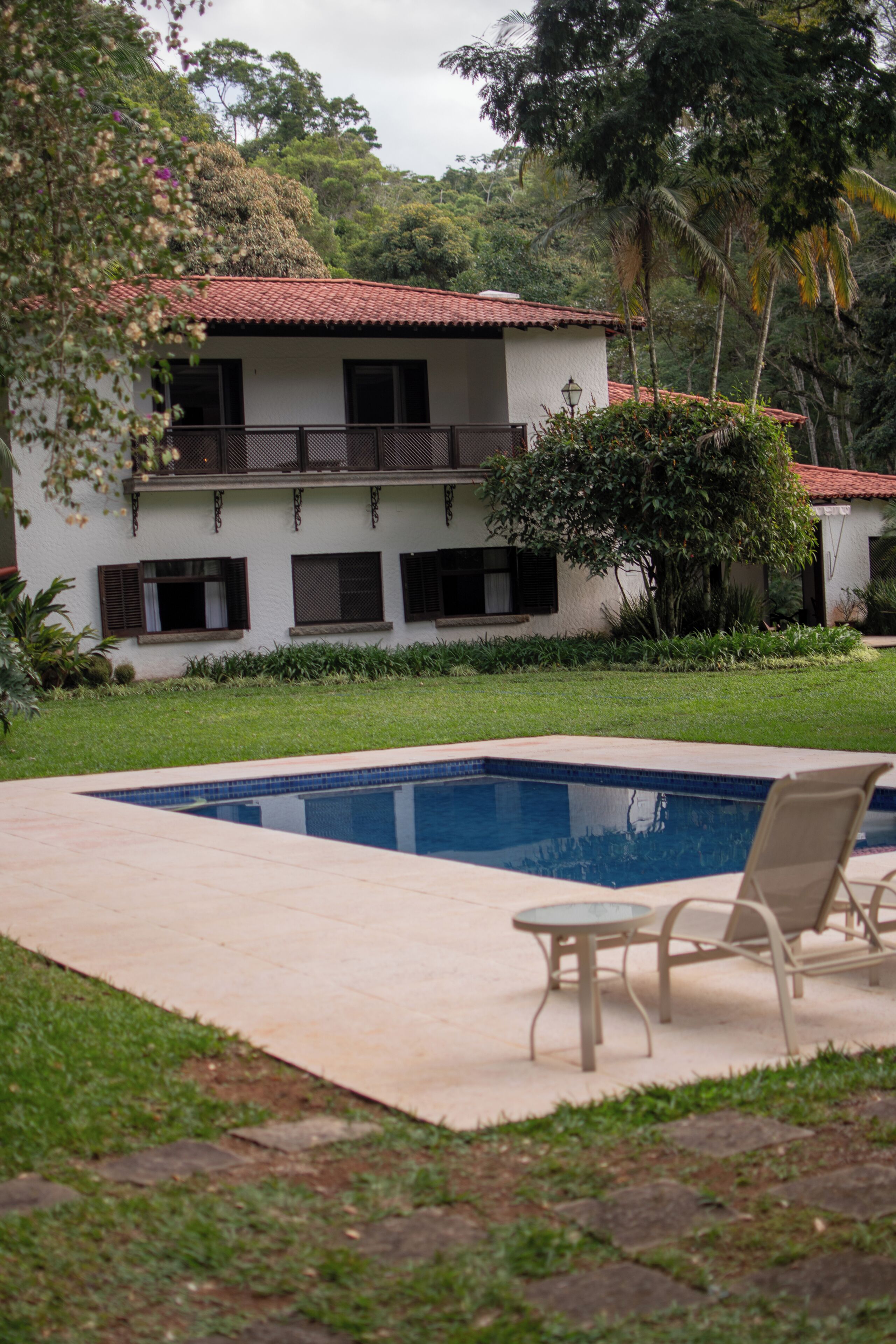 A pool with lounge chairs, a white building with a red roof, and surrounding greenery.