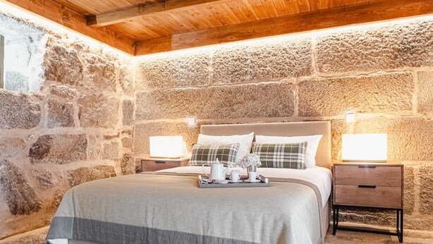 Une chambre avec un mur en pierre, un plafond en bois, un lit recouvert d’une couverture grise et blanche, deux tables de chevet avec des lampes, et un plateau contenant divers objets posé sur le lit.