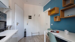 A compact room with a white door, a small window, a white table with a chair, and a blue wall with wooden shelves. - Italianway - San Calocero 29 B (Milano)