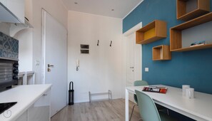 A compact room with a white door, a small window, a white table with a chair, and a blue wall with wooden shelves. - Italianway - San Calocero 29 B (Milano)