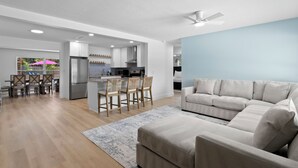 Un salon moderne avec un canapé d’angle, un coin repas avec des chaises en bois, une cuisine avec des meubles blancs et un ventilateur de plafond.