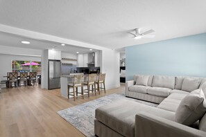 Un salon moderne avec un canapé d’angle, un coin repas avec des chaises en bois, une cuisine avec des meubles blancs et un ventilateur de plafond.