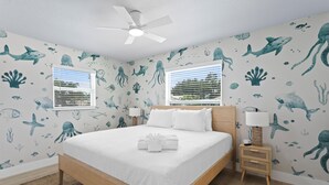 Une chambre à coucher comprenant un lit, deux tables de chevet, un ventilateur de plafond et des murs décorés d’autocollants représentant des motifs marins.