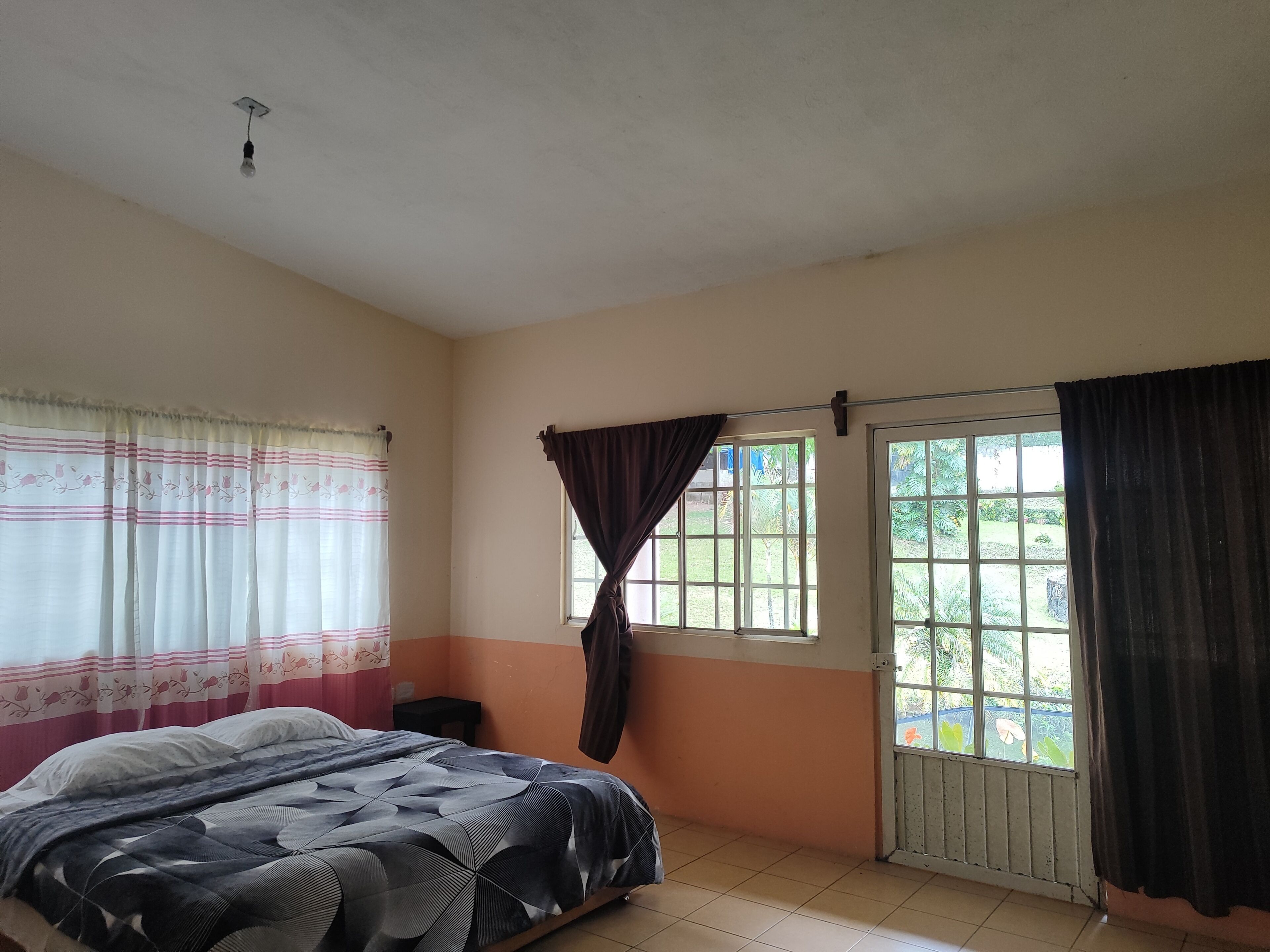 Un dormitorio con cama, ventana con cortinas, puerta y lámpara en el techo.