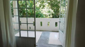 Puerta abierta que da a un balcón con vistas a árboles y una pared blanca con pequeñas ventanas.