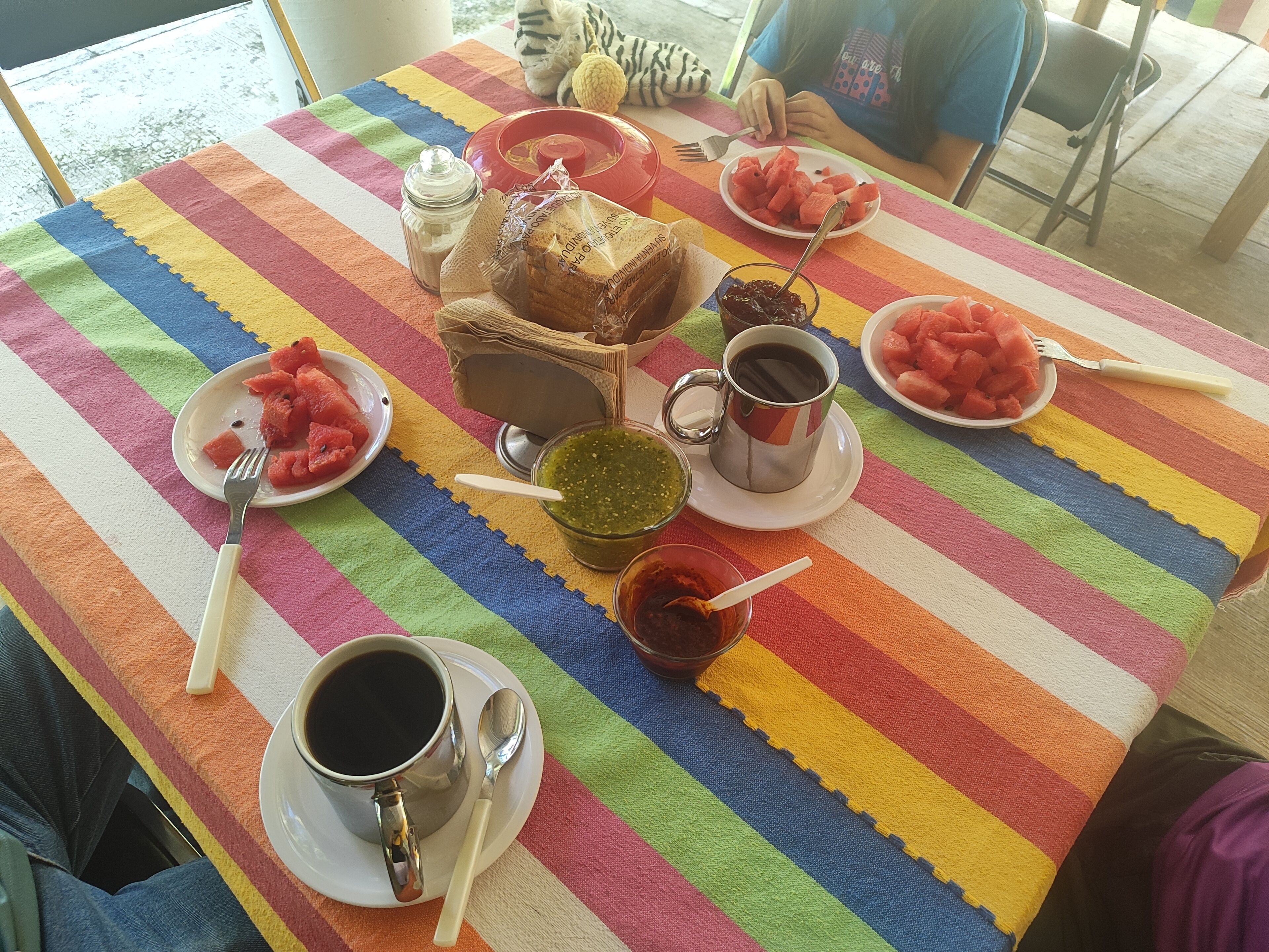 Una mesa con comida y utensilios, incluyendo platos de sandía, un tarro de mermelada y una taza de café.