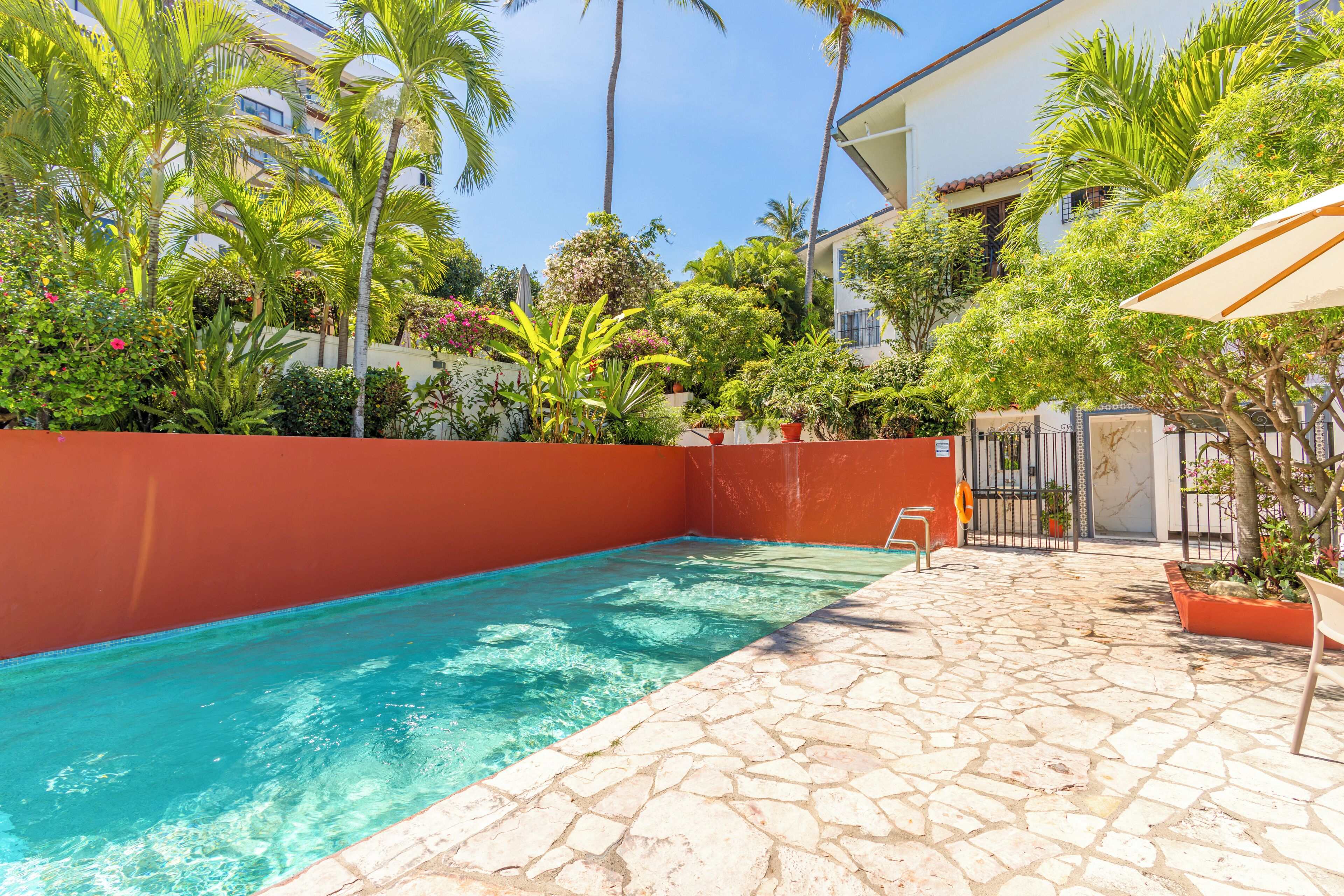 Une piscine avec une terrasse en pierre, entourée d’une végétation luxuriante et un bâtiment en arrière-plan.