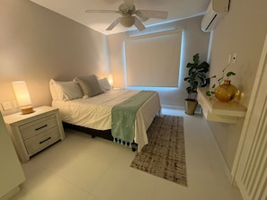 A bedroom with a bed, bedside table, lamp, ceiling fan, window, potted plant, and a small shelf with decorative items. - LOMA DEL MAR A19 PUERTO VALLARTA (Puerto Vallarta)